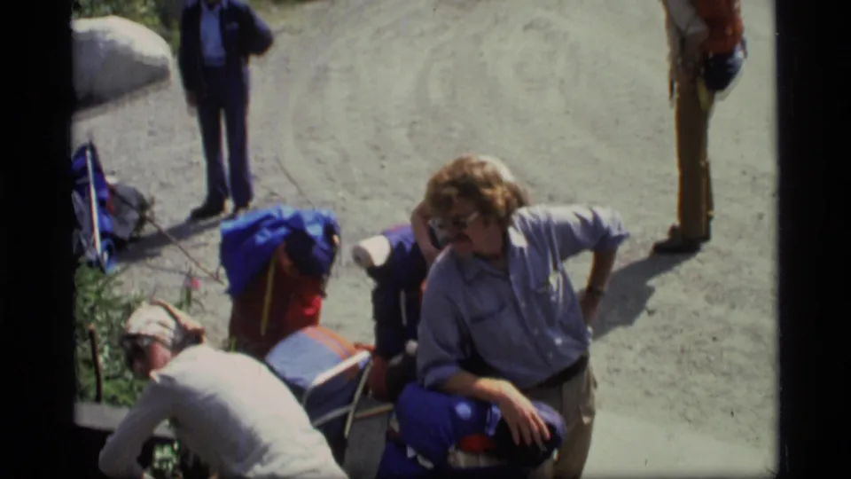 Home-movie frame from Glacier Bay in 1977 showing a casual group rest with backpacks, caps, and hikers on a gravel path.