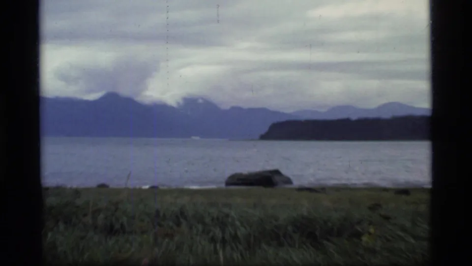 Glacier Bay, Alaska shoreline in 1977 with a large rock, grass, calm water, distant mountain silhouettes, and heavy cloud cover.
