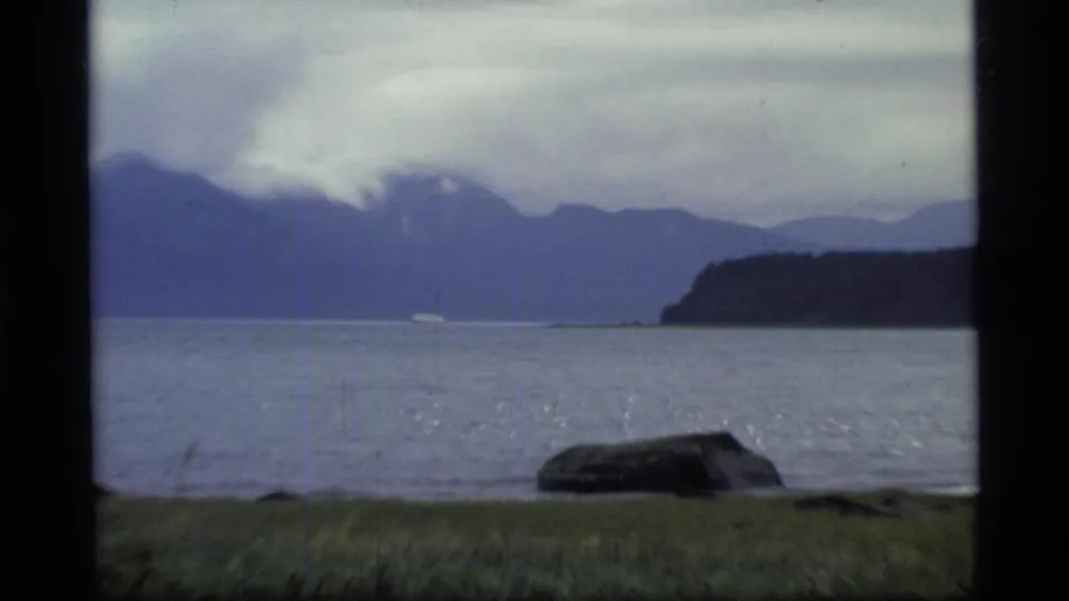 1977 Glacier Bay home-movie frame showing calm water, blue mountain ridges, cloudy sky, grassy shore, and a dark rock in the foreground.