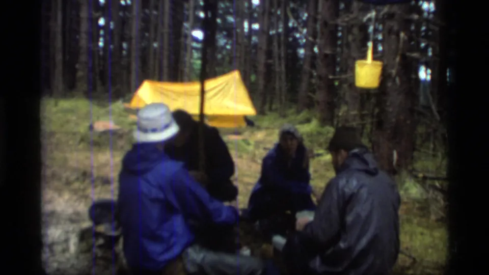 Friends in rain gear sit at a wooded campsite near a yellow tent in 1977 Glacier Bay home-movie footage.