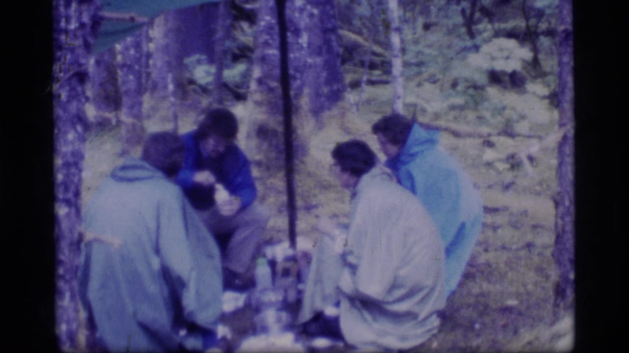 Campers sit under a tarp in a wooded campsite, gathered around food and gear.