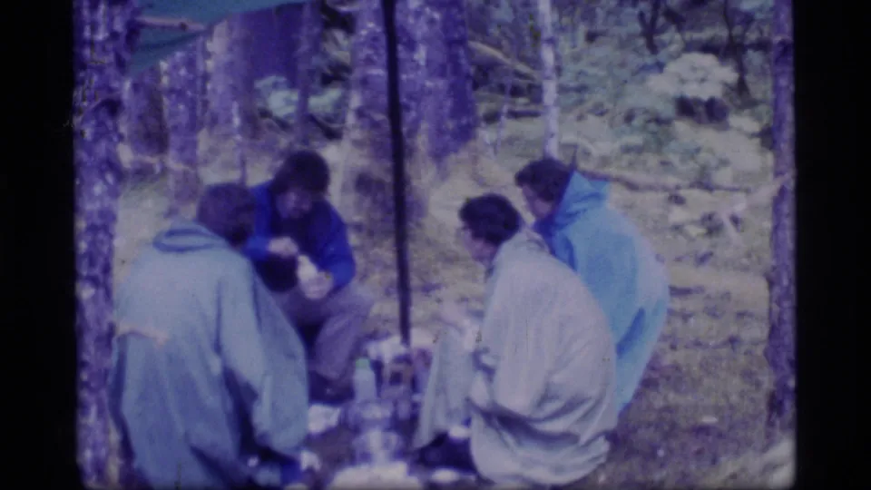 1977 Glacier Bay home-movie frame showing campers in rain gear seated under cover in a forest campsite.