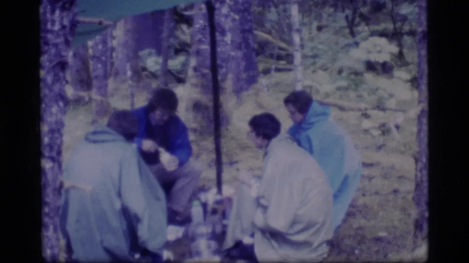 Campers in Glacier Bay, Alaska, appear seated under a tarp or canopy during a rainy 1977 forest camping scene.