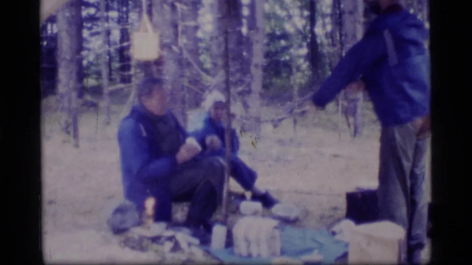 1977 8mm frame of campers in a Glacier Bay forest campsite, with one standing figure near seated people and gear.