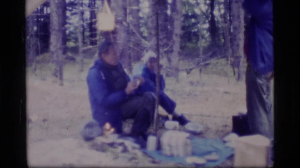 Glacier Bay camping home-movie frame with seated campers, a partial standing figure, rain gear and campsite supplies.