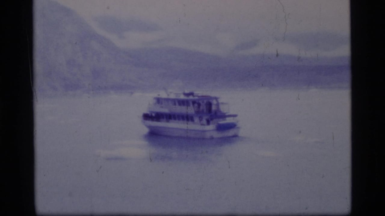 1977 8mm home-movie frame of a ferry crossing calm water in Glacier Bay, Alaska, with soft blue mountain scenery behind it.