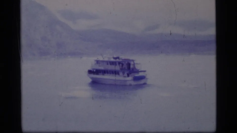 1977 8mm home-movie frame of a ferry crossing calm water in Glacier Bay, Alaska, with soft blue mountain scenery behind it.