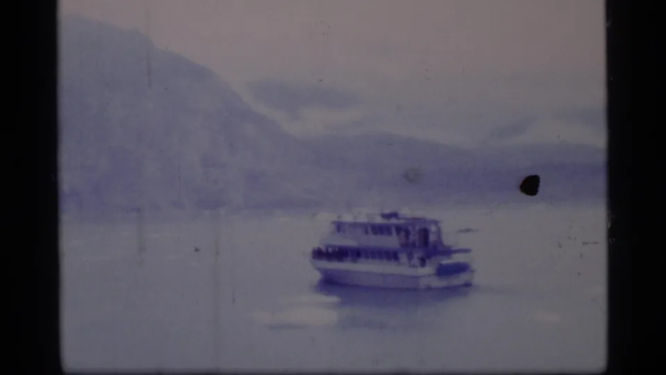 Blue-tinted 8mm frame showing a ferry on Glacier Bay water, backed by distant mountain slopes and visible film wear.