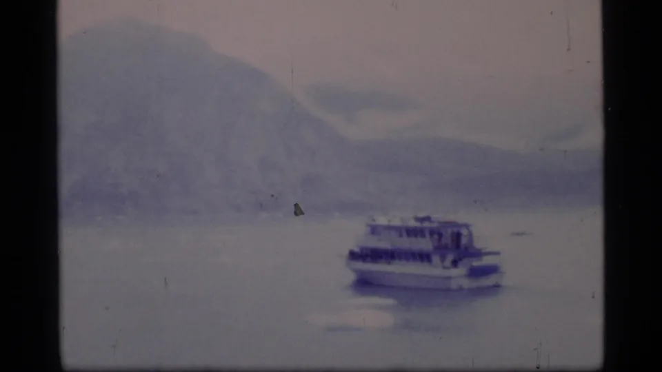 Soft 1977 8mm view of a ferry moving across Glacier Bay with pale mountains, calm water, and visible home-movie film texture.