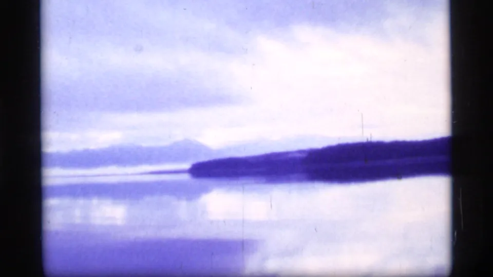 Glacier Bay boat-view frame showing calm water, a dark shoreline, distant mountains, and a cloudy sky in faded 8mm color.