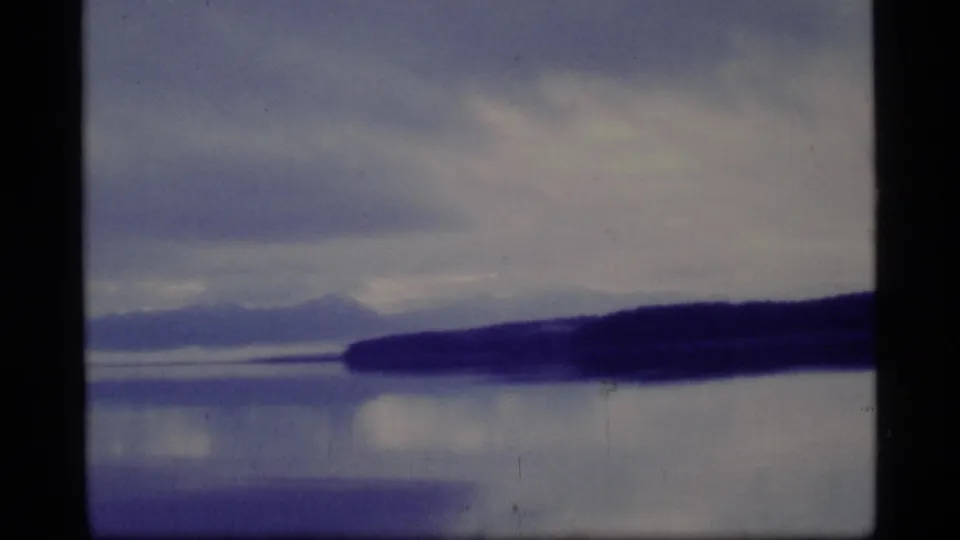 1977 Glacier Bay 8mm frame with open water, a low dark shoreline, hazy mountain forms, and muted overcast light.