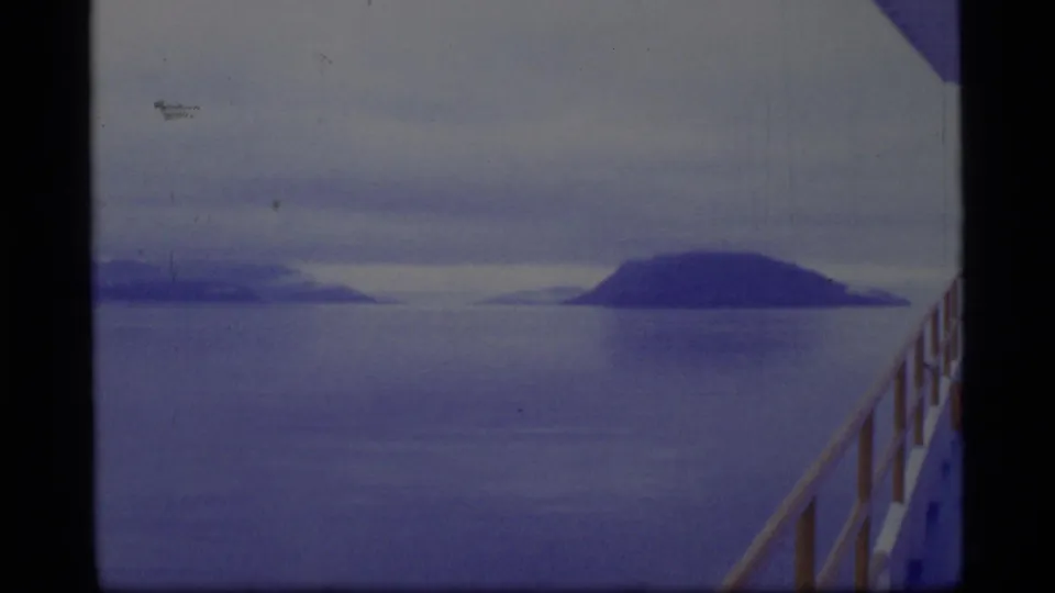 Glacier Bay travel frame from a boat, with open water, distant islands, cloudy sky, and a railing visible along the right side.