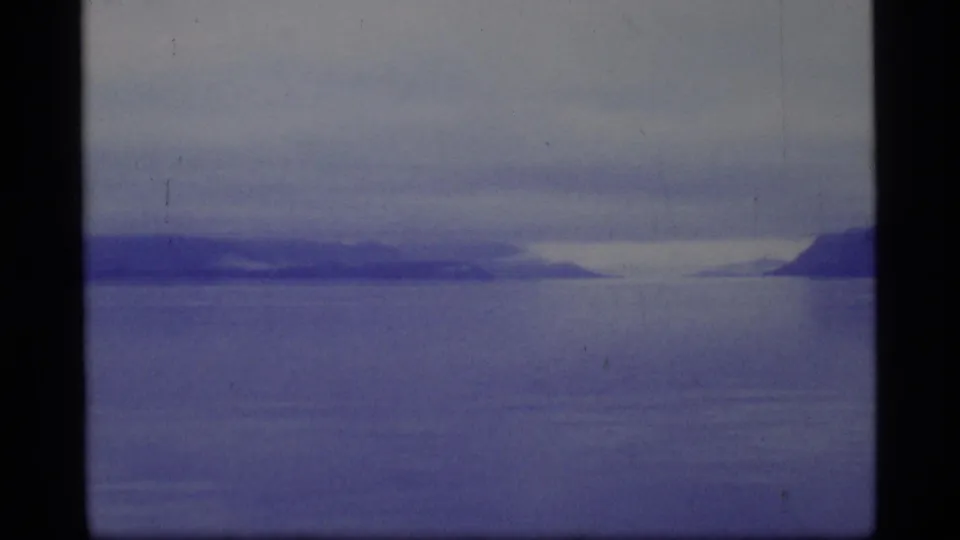 A wide 1977 home-movie view of Glacier Bay water with muted blue tones, cloudy sky, and distant mountain shapes along the horizon.