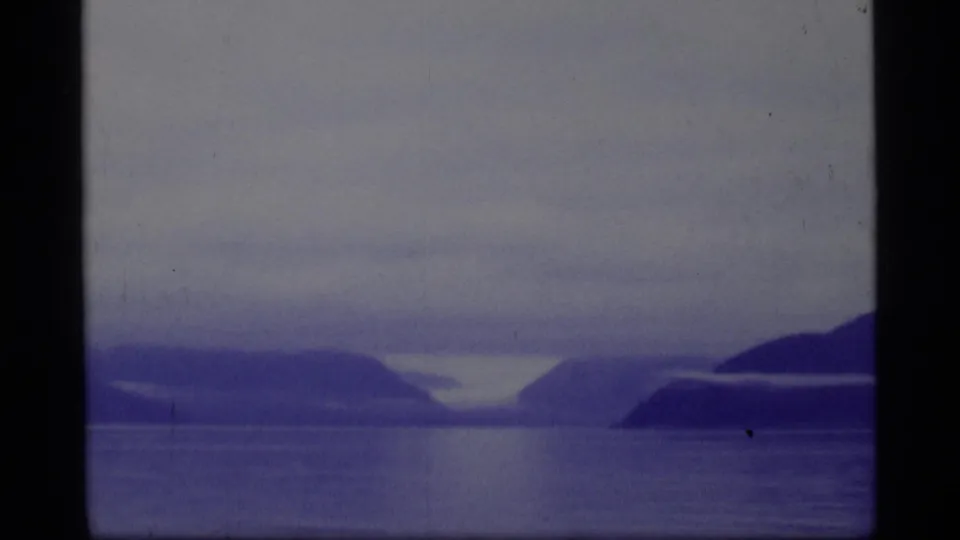 Glacier Bay landscape on 8mm film, with quiet water, layered mountain forms, and a dark shoreline mass at right.