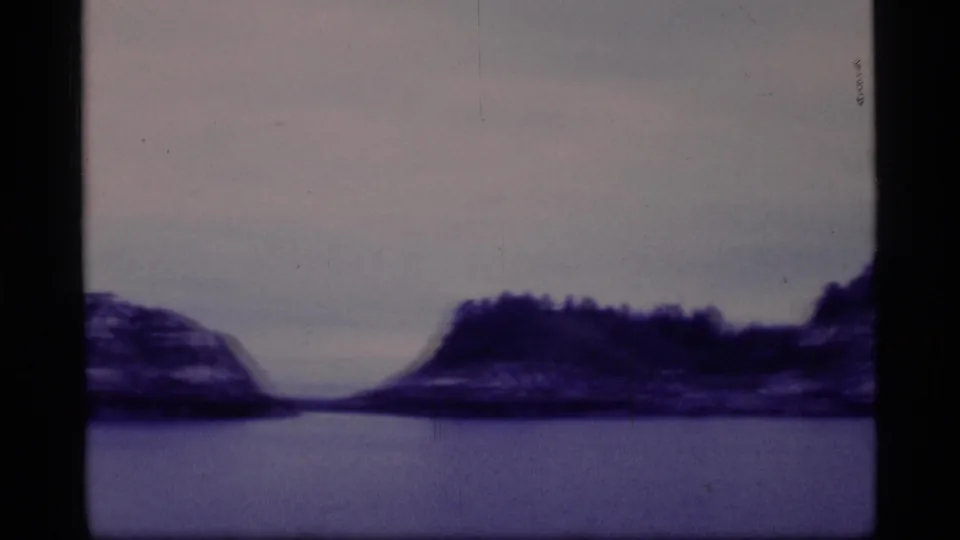 Glacier Bay shoreline with dark trees, low rocky landforms, and smooth water, recorded in a soft 8mm home-movie frame.