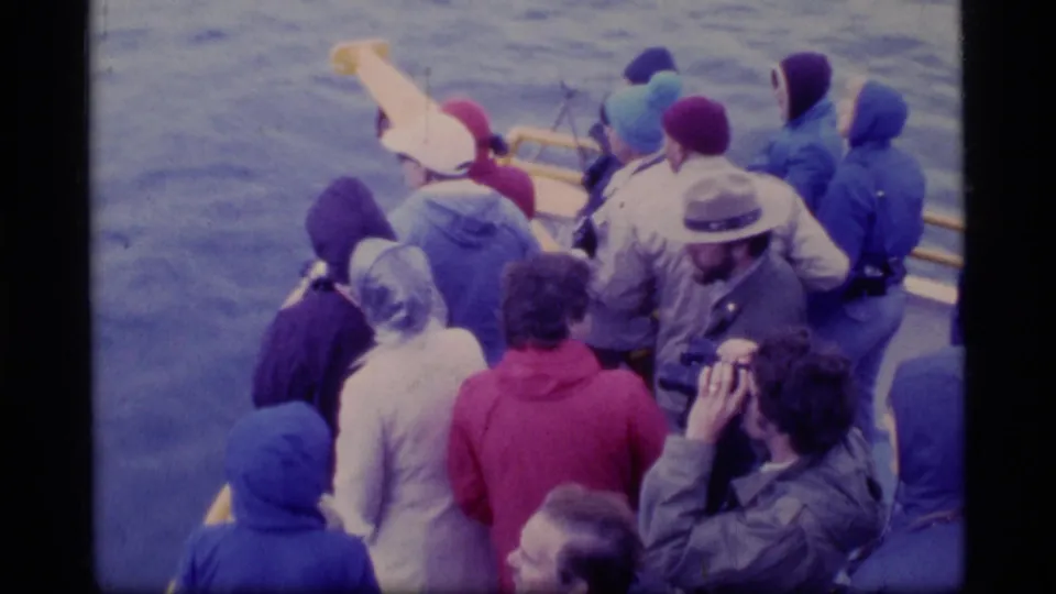 Tourists bundled in coats crowd the boat rail in Glacier Bay, with one passenger lifting a camera toward the view.
