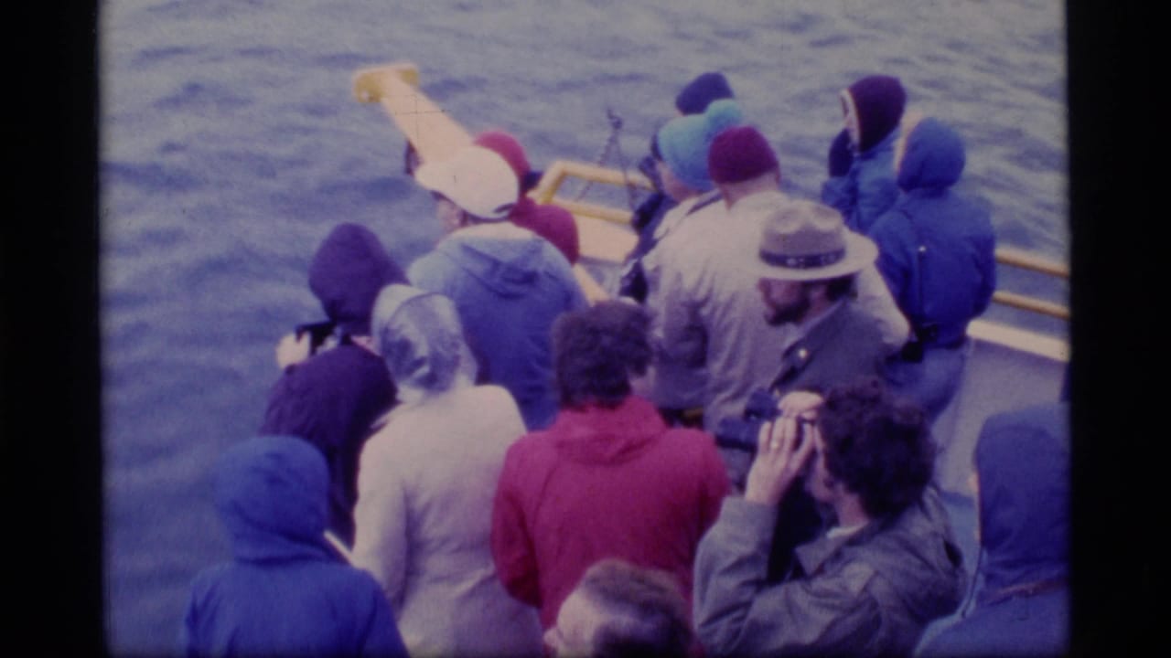 Passengers in coats gather at a boat rail over gray-blue water.