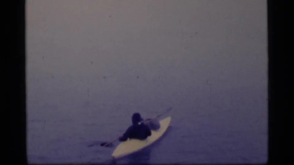 A distant kayaker paddles across calm Glacier Bay water in a 1977 8mm home-movie frame, with no face detail visible.