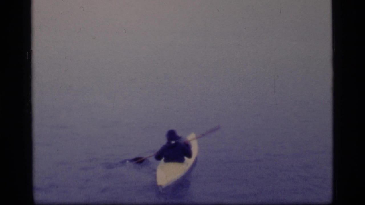 In 1977 Glacier Bay home-movie footage, a distant kayaker in dark clothing paddles a pale kayak across calm water.
