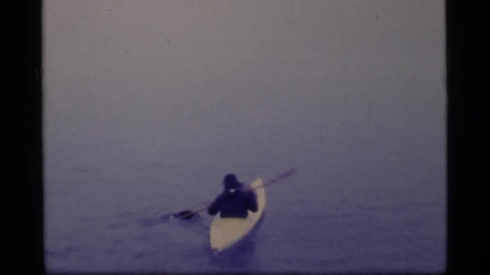 In 1977 Glacier Bay home-movie footage, a distant kayaker in dark clothing paddles a pale kayak across calm water.