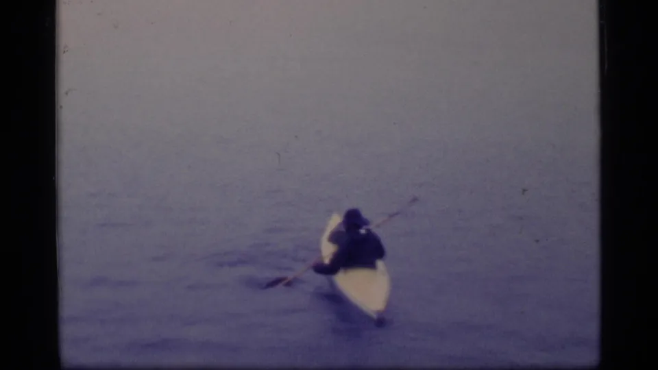 A distant person in a dark jacket paddles a light-colored kayak on the calm waters of Glacier Bay, Alaska.