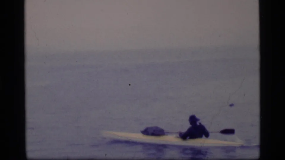 Side-on view of the lone kayaker in Glacier Bay, with a pale kayak crossing a muted blue-gray water surface.