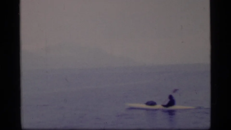 A distant kayaker paddles across calm water in Glacier Bay, Alaska, with soft 1970s 8mm color and mountains behind.