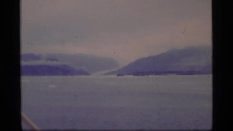 Glacier Bay water and hazy mountain slopes appear under cloud cover in a soft, grainy 1977 home-movie frame.
