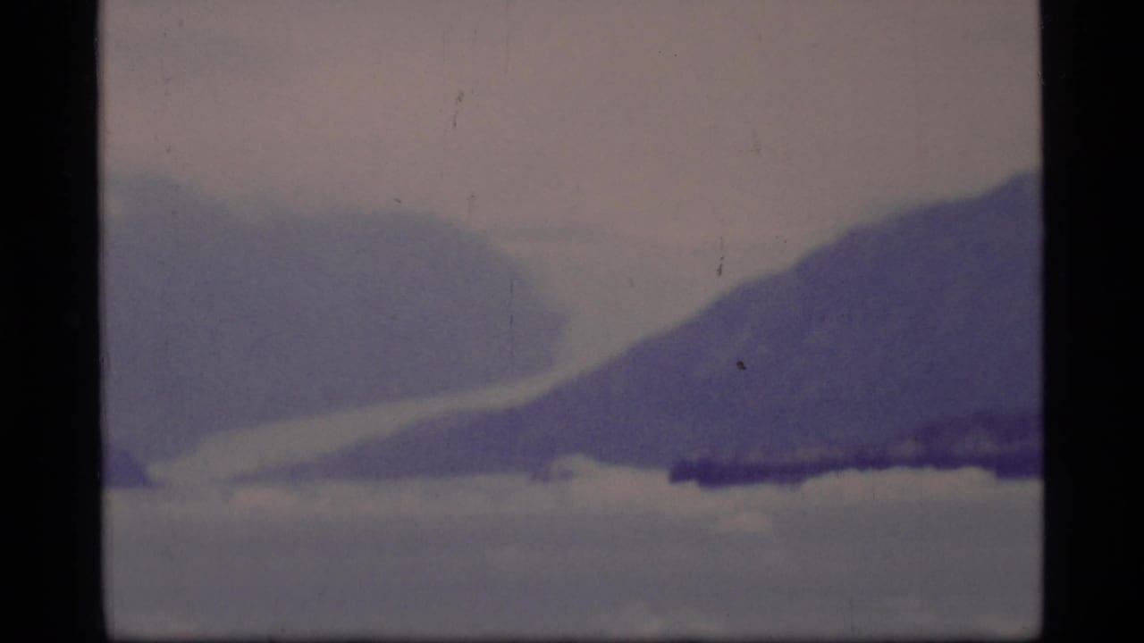 1977 8mm Glacier Bay view of a hazy mountain shoreline across cold water, tinted blue-purple by aged film.