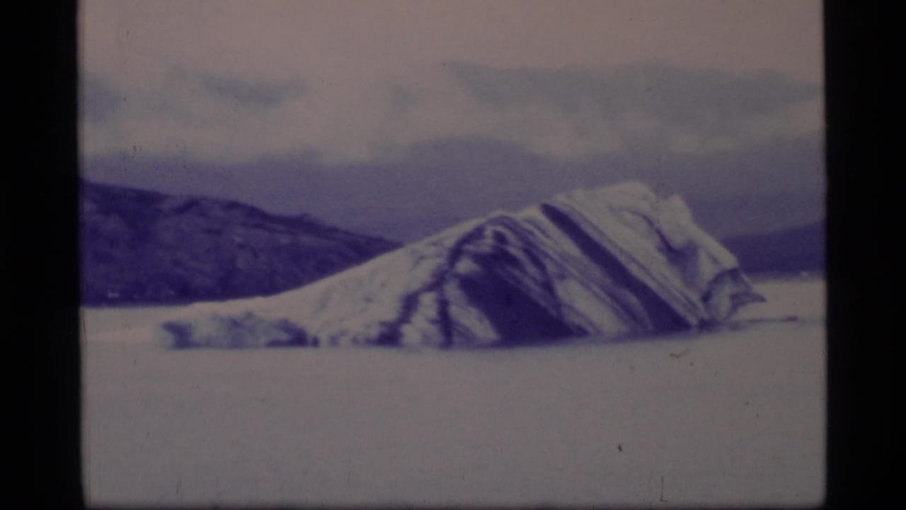 Large striated iceberg floating before dark ridges and cloudy sky.
