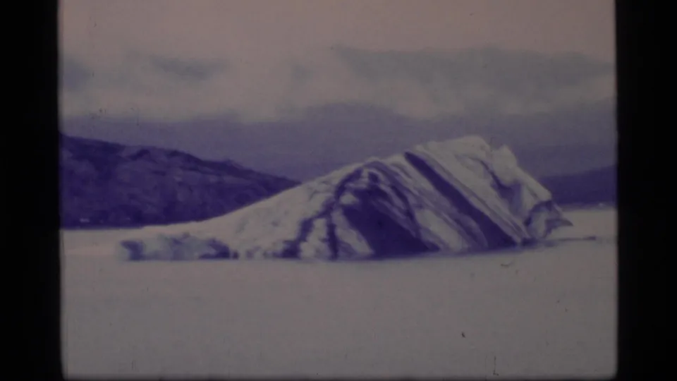 1977 Glacier Bay frame showing a large striated iceberg in cold water with dark mountain ridges behind it.