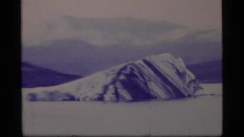 1977 8mm view of a glacier-like iceberg form resting on pale water in Glacier Bay with low clouds overhead.