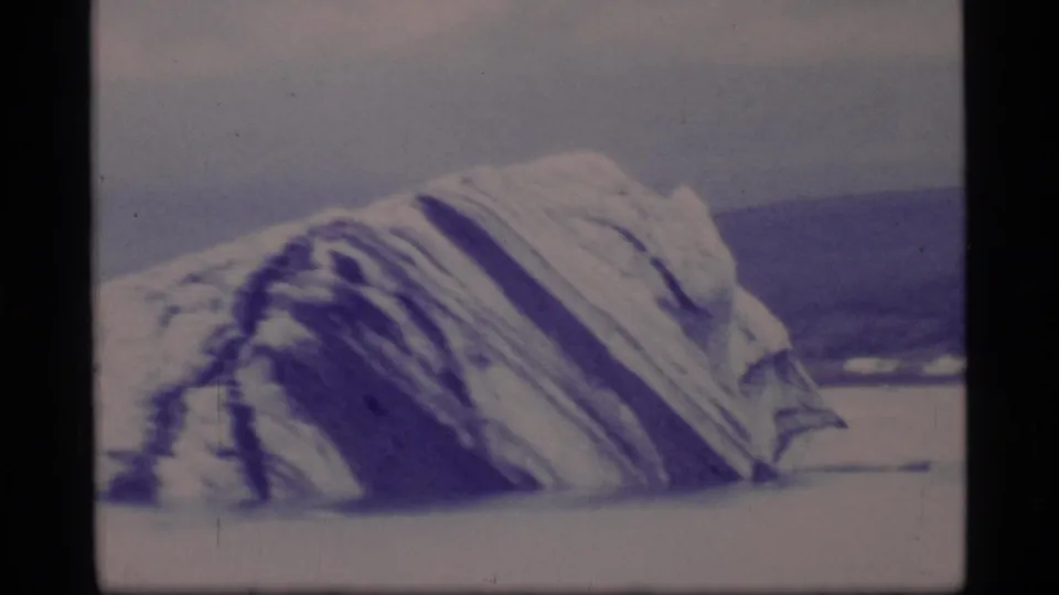 Closer 1977 Glacier Bay 8mm frame of a ridged iceberg, with blue-purple shadowing and distant shoreline beyond.