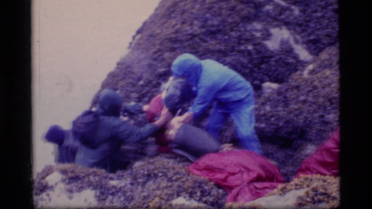 Hikers in hooded rain gear assist each other on a rocky slope.
