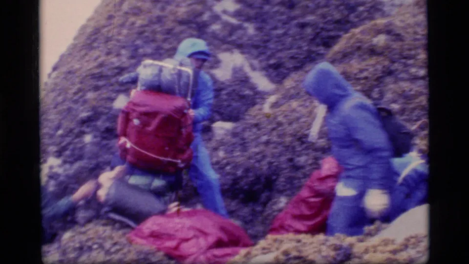 Glacier Bay hikers in blue rainwear and backpacks navigate slick rock, with red packs visible in the foreground.