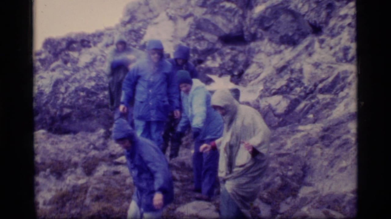 1977 Glacier Bay home-movie frame showing hikers in blue and light rain gear navigating a wet, rocky mountainside.