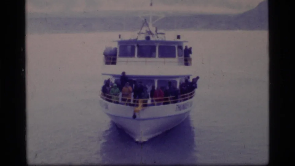 Glacier Bay travel frame showing a passenger boat approaching the camera on calm water, with people visible along the front deck.