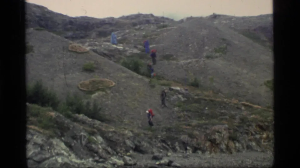 Wide 1977 Glacier Bay home-movie view of hikers crossing a gray, rocky hillside with low vegetation and rounded terrain.