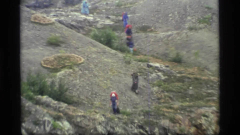 Glacier Bay home-movie frame showing several hikers in colorful rain gear spaced along a rocky path in sloped terrain.