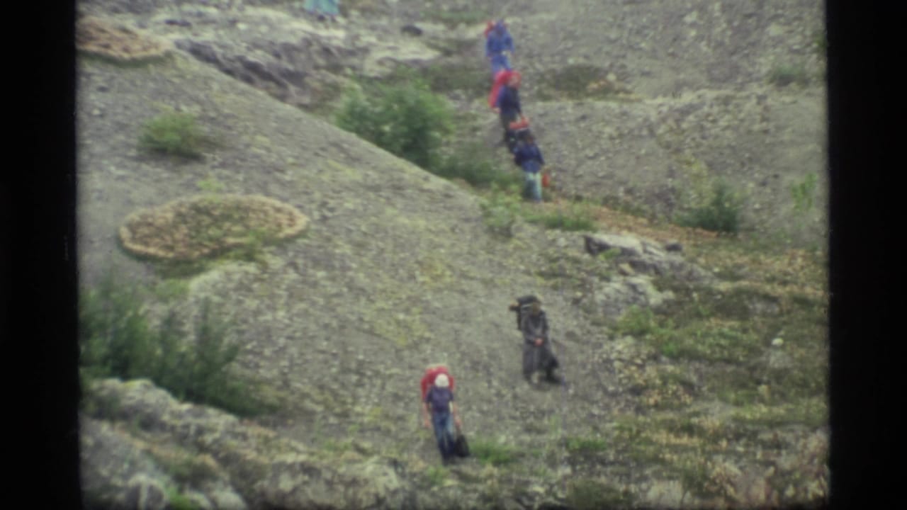 1977 Glacier Bay home-movie frame showing distant hikers with packs moving down a rocky Alaskan slope.