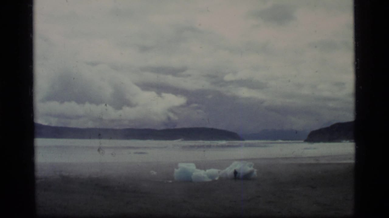 1977 Glacier Bay home-movie frame showing a gray shoreline, blue-white ice pieces, dark mountains, and low clouds.
