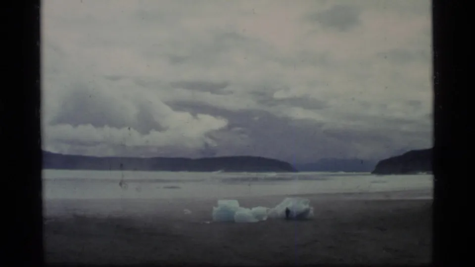 1977 Glacier Bay home-movie frame showing a gray shoreline, blue-white ice pieces, dark mountains, and low clouds.