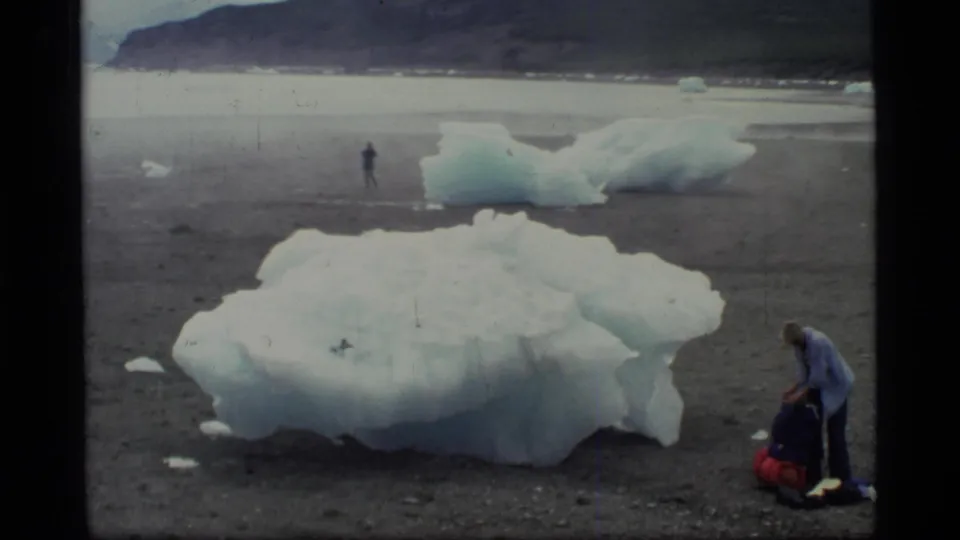 1977 Glacier Bay frame with blue-white ice blocks on gravel, a distant figure, and a backpacker adjusting gear at right.
