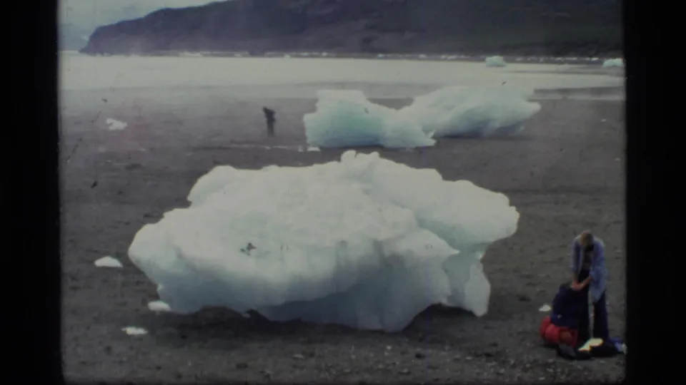 1977 Glacier Bay frame showing a backpacker bending over gear beside a large blue-white ice mass on a barren plain.