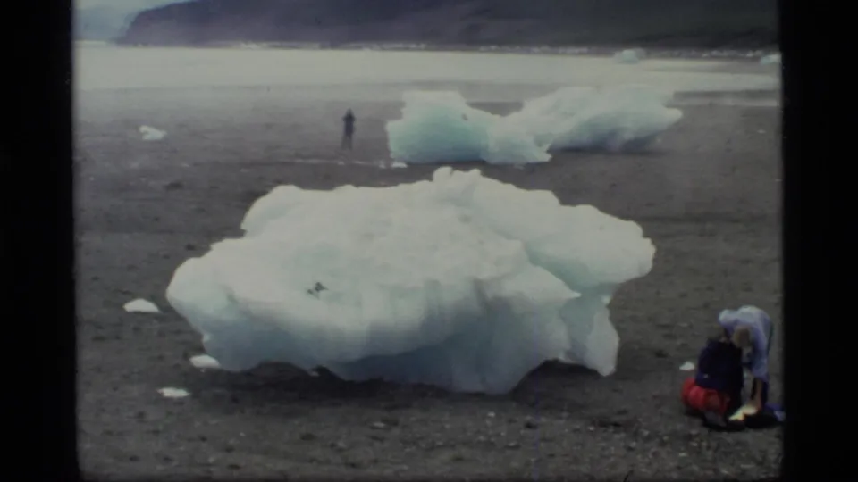 1977 Glacier Bay frame with a large blue-white ice formation, a backpacker at right, and another person standing far back.