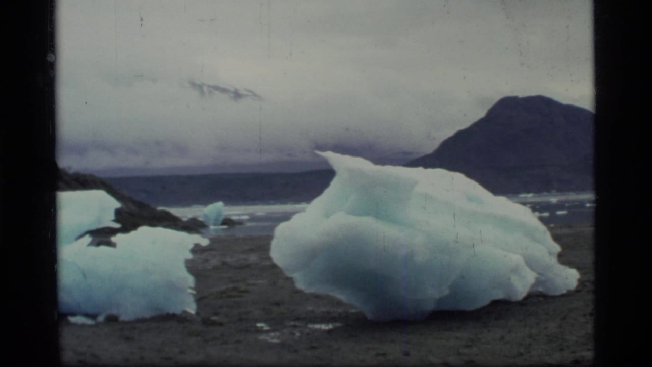 1977 8mm Glacier Bay view of a stranded iceberg on a rocky shoreline under a gray overcast sky.