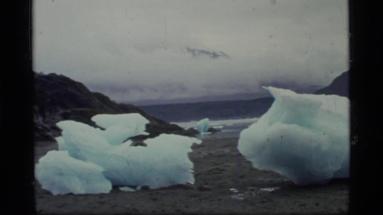 Blue ice chunks sit along a rocky shoreline beside gray bay water and distant hills.
