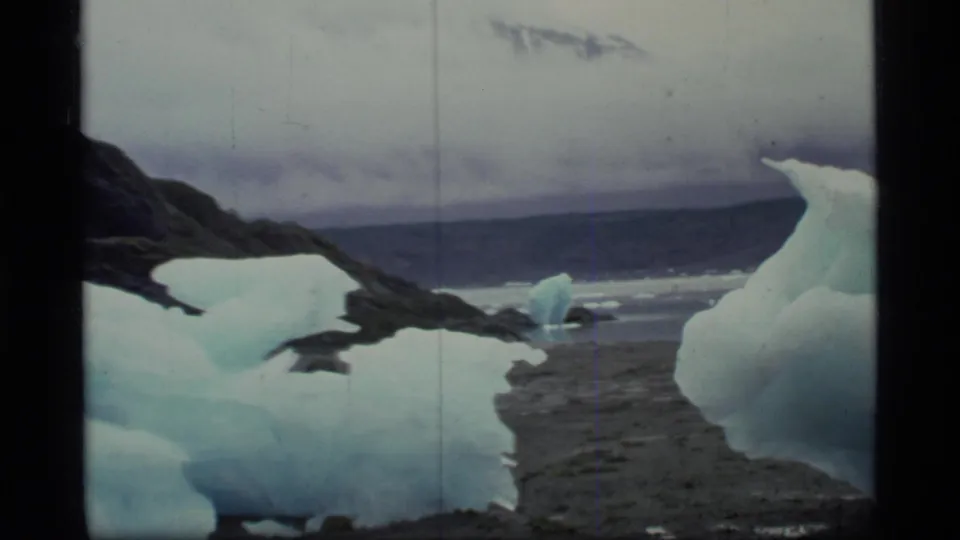 Home-movie frame of foreground glacier ice on a Glacier Bay beach with distant water and low clouds.