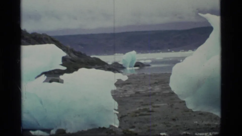 1977 8mm view across Glacier Bay shoreline ice toward a small iceberg, gray water, and dark mountain ridge.