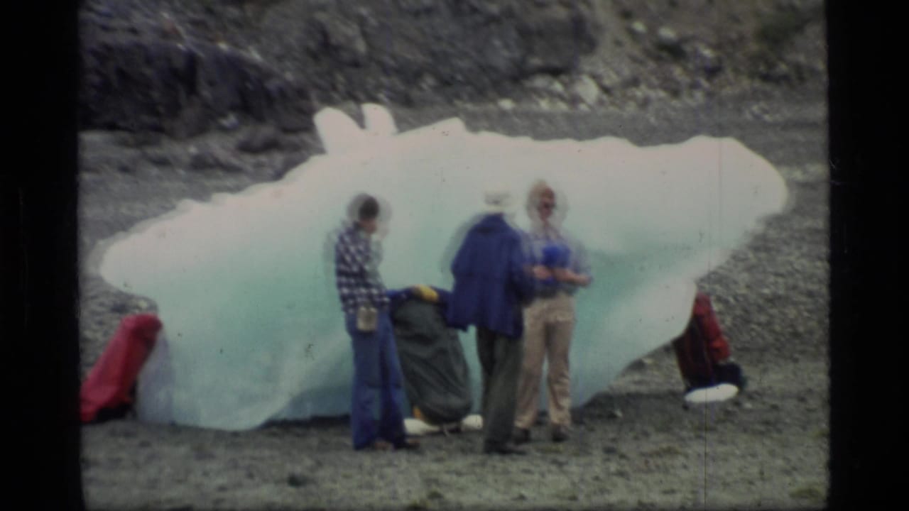 In Glacier Bay, Alaska, three adults in cold-weather clothing examine a large translucent ice block on a rocky beach.